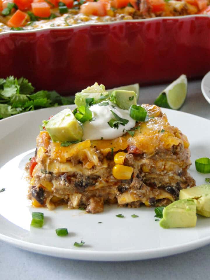 Side view of mexican lasagna slice topped with dollop of sour cream, diced avocado and cilantro leaves on white round plate in front of casserole dish.