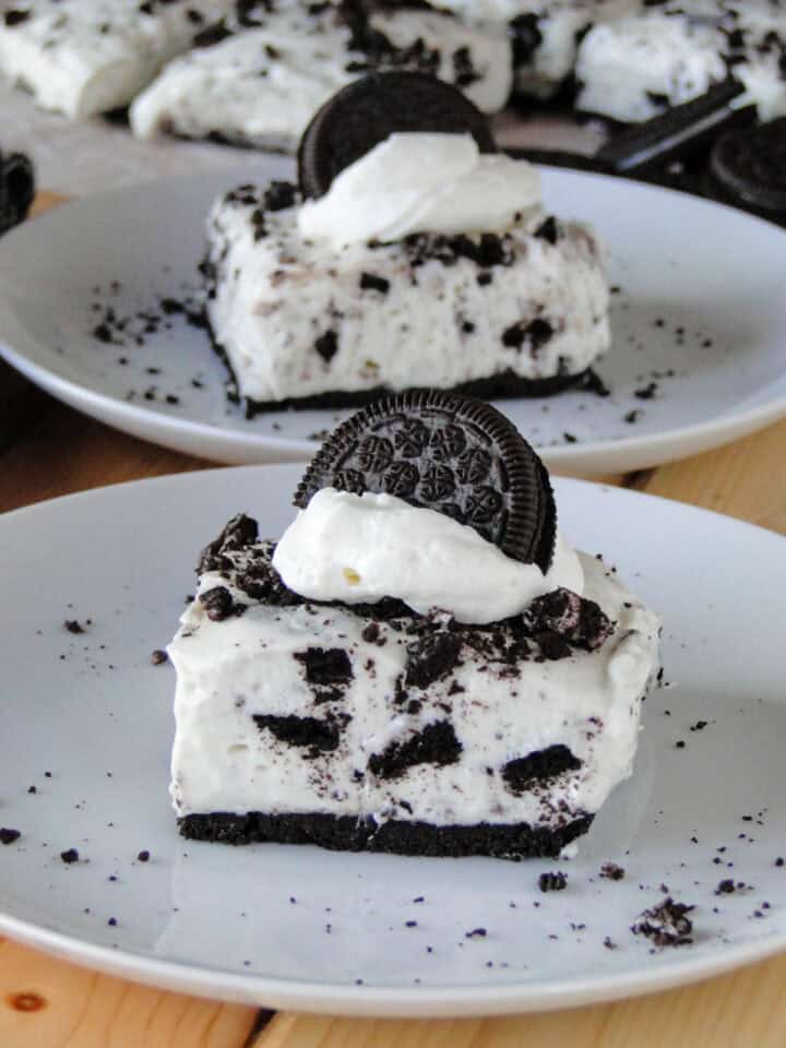 Side view of slice of no bake cookies and cream cheesecake bar on white round plate in front of another slice on white round plate.