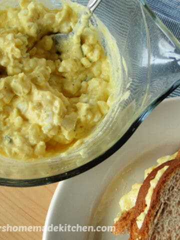 Classic Egg Salad in clear bowl with spoon next to sandwich.