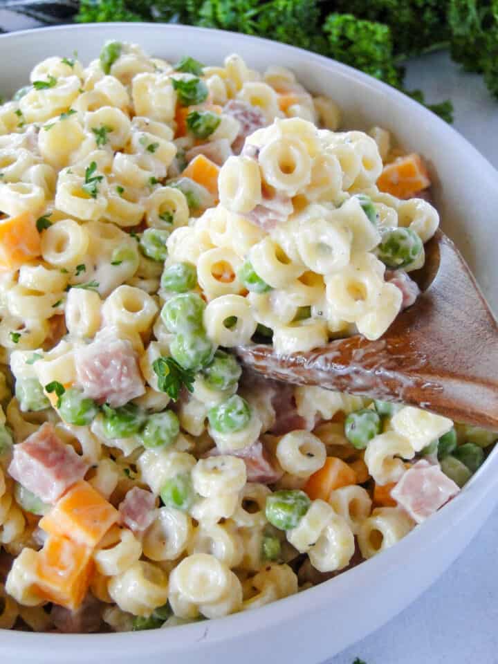 Side view of ham pasta salad in white round bowl with scoop on wooden serving spoon.