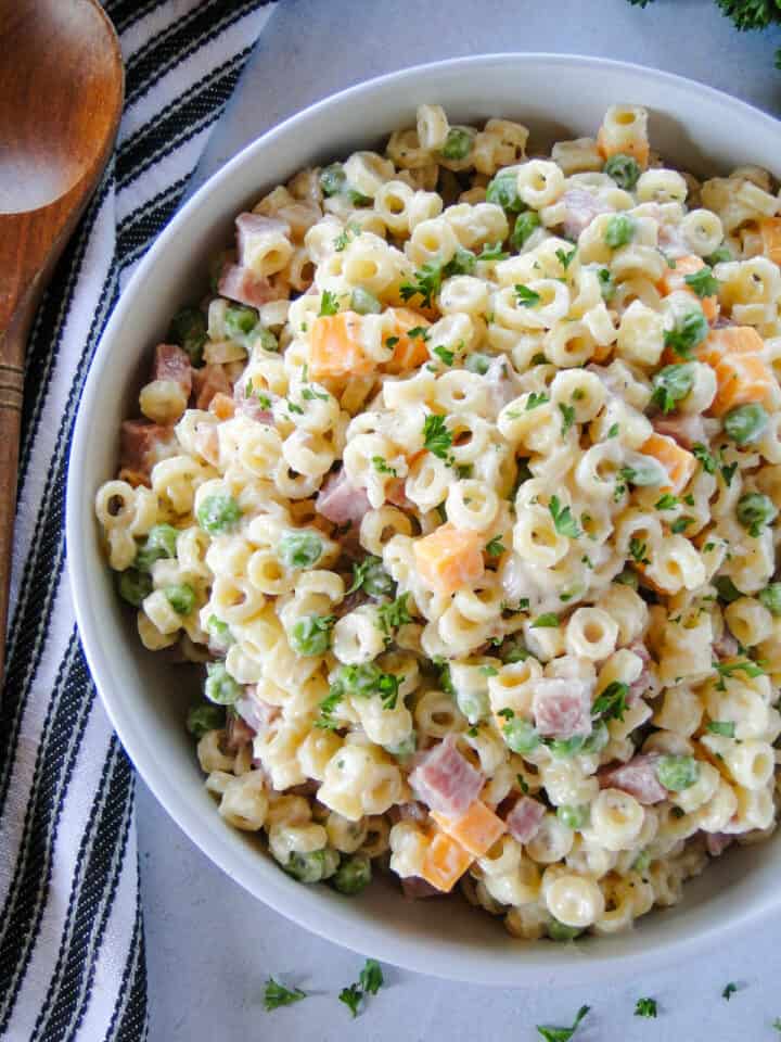 Ham pasta salad in white round bowl with wood serving spoon on the left side.