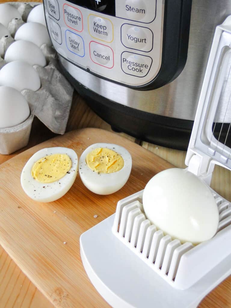 Instant Pot hard boiled eggs in carton next to instant pot, an egg sliced in half on board and an egg on the egg slicer. 