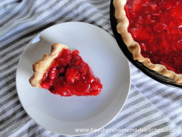 Top View of Fresh Strawberry Pie Slice on white round plate sitting next to full pie. 