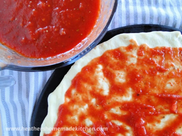Homemade Pizza Sauce No Cook in glass bowl next to pizza dough with sauce on top on pizza stone.