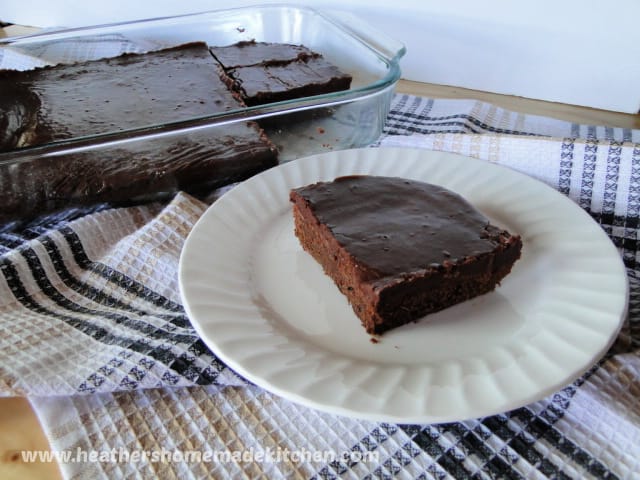 A slice of Zucchini Brownies with Fast Frosting on round white plate with dish of brownies behind.