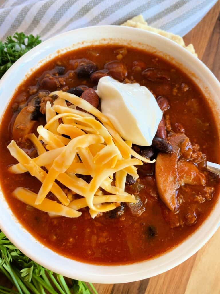 Top view of 3 bean chili in white bowl with spoon and topped with sour cream and yellow shredded cheese.