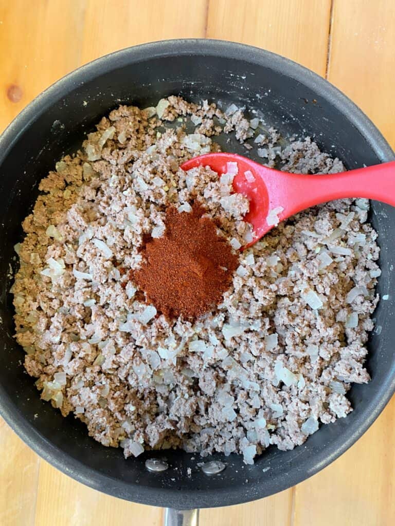 Cooked ground beef in pan with chili powder and red spoon.