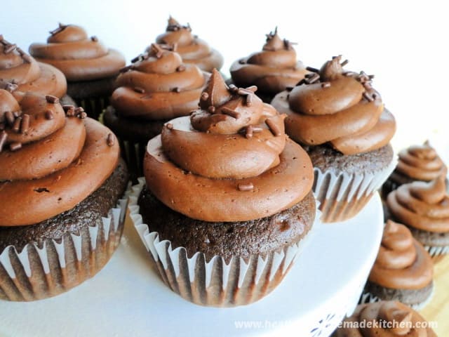 Homemade Chocolate Cupcakes with chocolate buttercream frosting and chocolate sprinkles on round white cake stand.