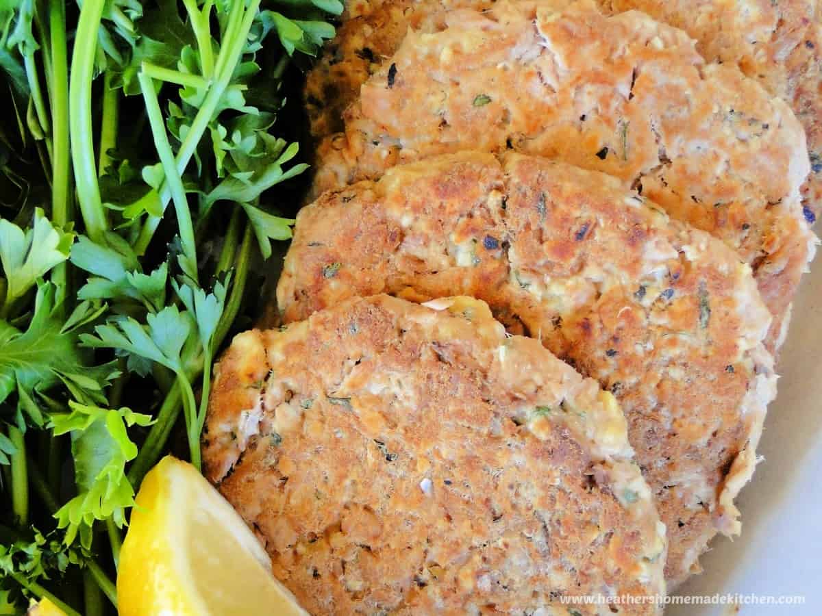 Healthy basil tuna cakes spread on white platter with parsley and lemon wedges.