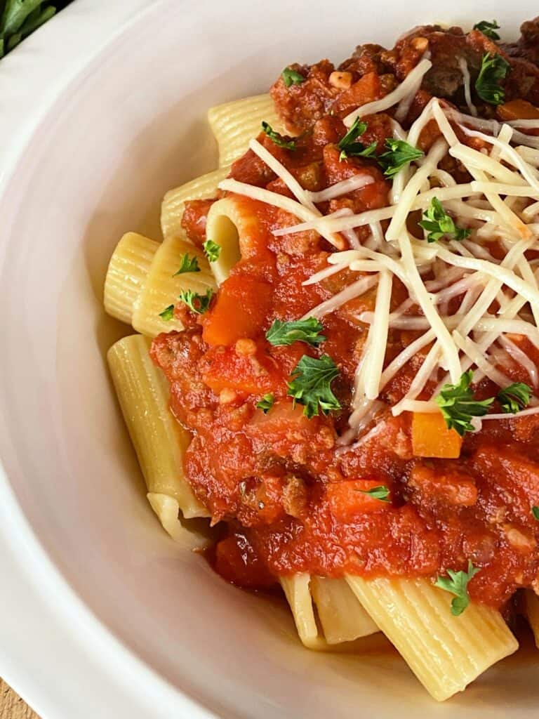 Close up view of crock pot beef bolognese over rigatoni topped with shredded parmesan and parsley pasta in white bowl. 