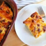 Best ever egg bake slice on white round plate with fork next to casserole dish of egg bake.