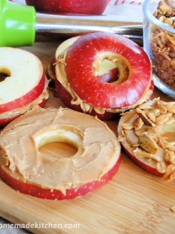 Peanut Butter Apple sandwiches with granola on board with apple slice and peanut butter and bowl of granola.