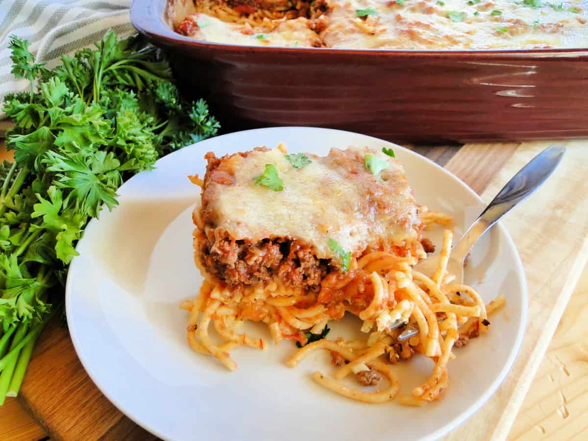 Slice of cheesy spaghetti bake on white round plate with bite on fork.