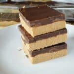 No bake peanut butter bars stacked 3 high on white round plate in front of glass baking dish with more bars.