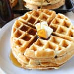 Homemade belgian waffles stacked on white round plate with pats of butter and getting a drizzle of maple syrup.