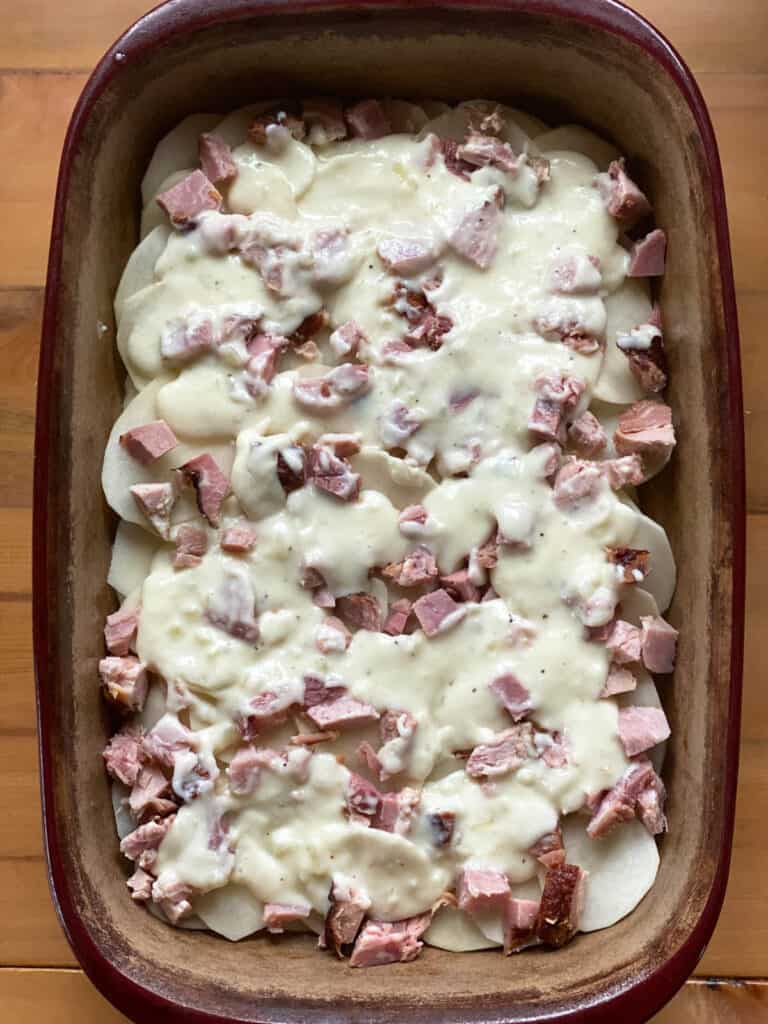 Béchamel sauce poured over top of ham and sliced potatoes in casserole dish.