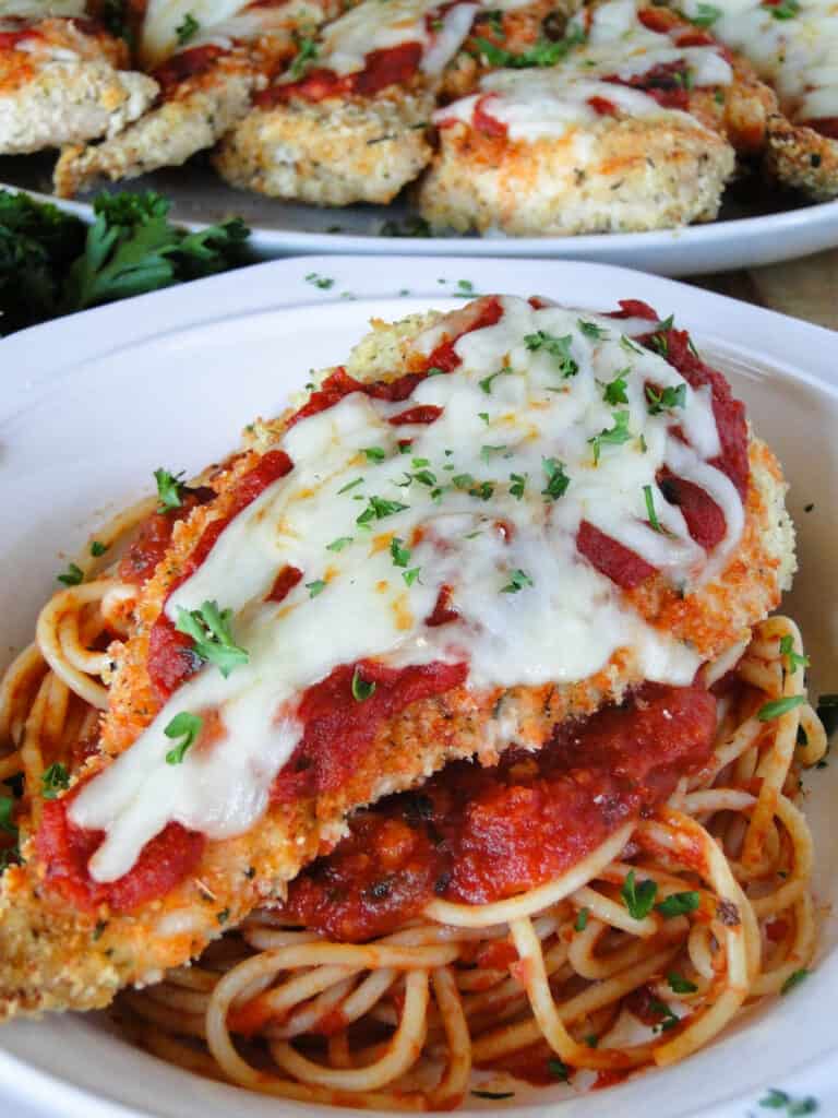 Close up view of baked chicken parmesan on top of spaghetti noodles in a white round pasta bowl.