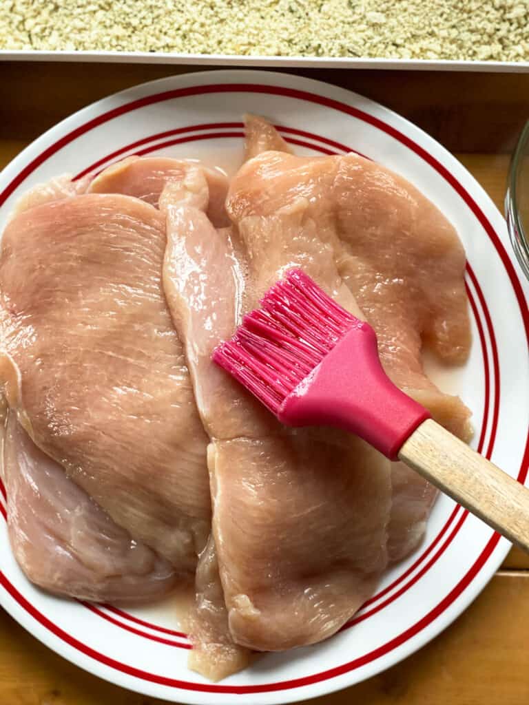 Chicken breast cutlets being brushed with beaten egg white.