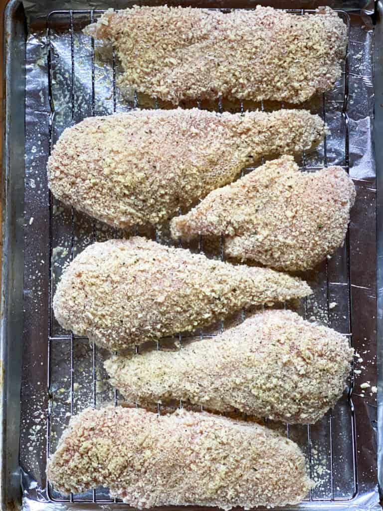 Chicken breasts coated with breading and sprayed with olive oil on baking rack inside a baking sheet.
