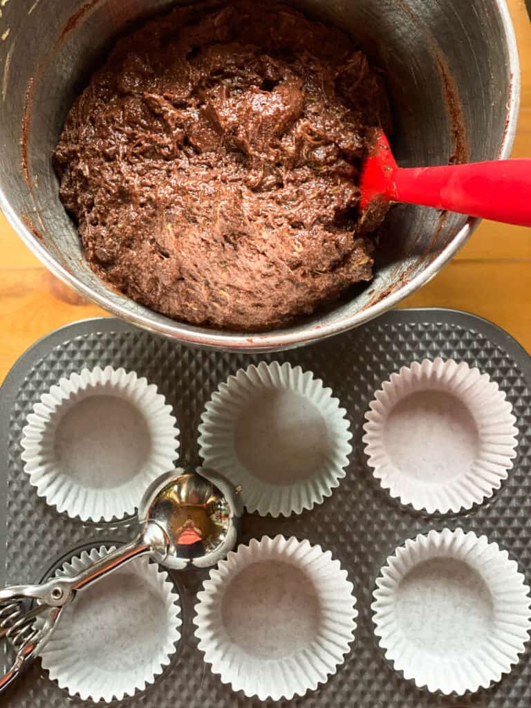 Mixed muffin batter in stand mixer bowl with paper lined muffin pan and scoop.