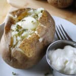 Instant pot baked potato on white round plate with pat of butter and parsley on top and side of sour cream on plate with a fork.