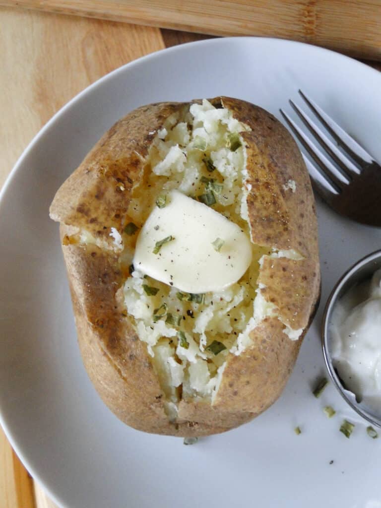 Top view of instant pot potato on white round plate with pat of butter and chives on top and sour cream and fork on plate.