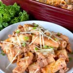 Baked rigatoni casserole on white round plate in front of red casserole dish.