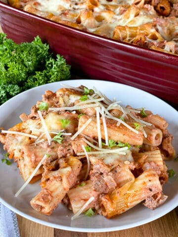 Baked rigatoni casserole on white round plate in front of red casserole dish.