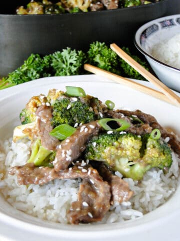 White bowl of rice topped with beef and broccoli stir fry and garnished with sesame seeds and green onions with chop sticks.