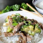 White bowl of rice topped with beef and broccoli stir fry and garnished with sesame seeds and green onions with chop sticks.