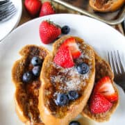 Top view Classic French toast piled 3 high on white round plate topped with blueberries, strawberries, melted butter and maple syrup.