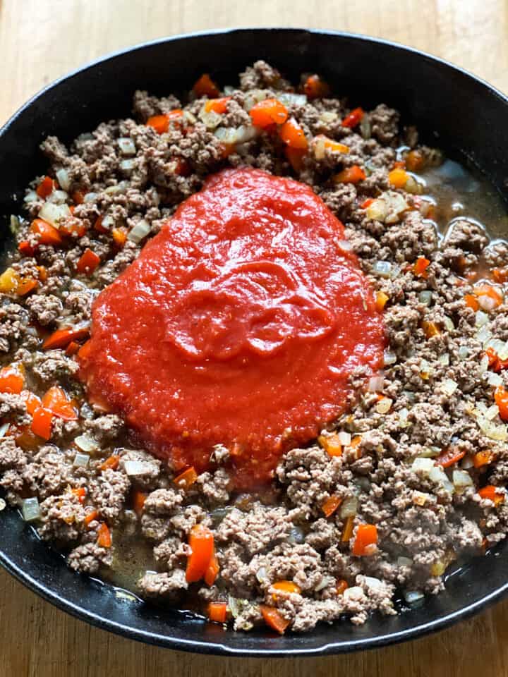 Broth and tomatoes added to cooked seasoned beef and veggies in cast iron pan.