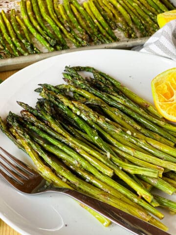 Roasted asparagus piled on white round plate with lemon half and fork.