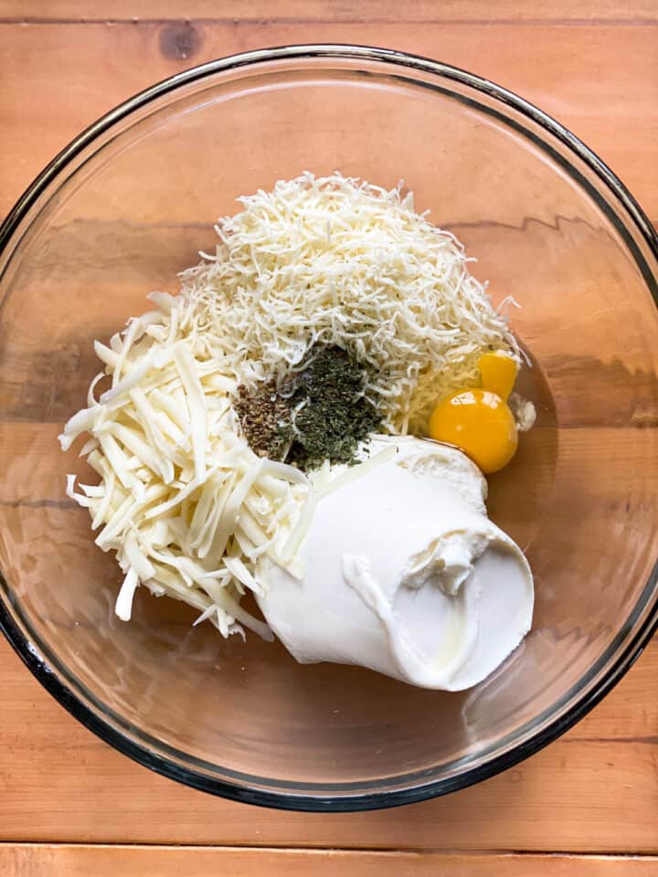 Shredded cheese, ricotta cheese, eggs and seasonings added to large glass bowl.