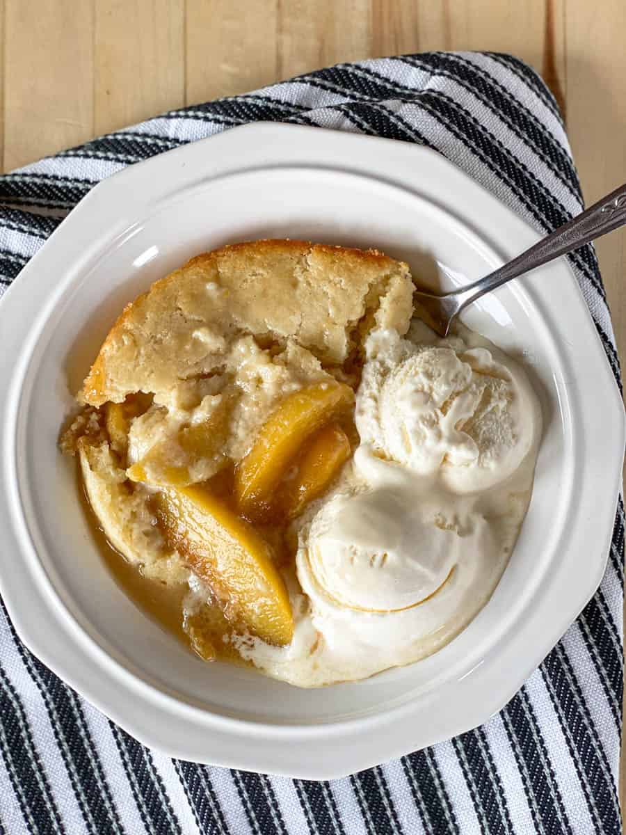 Top view of peach cobbler in white bowl with 2 scoops of vanilla ice cream and spoon.