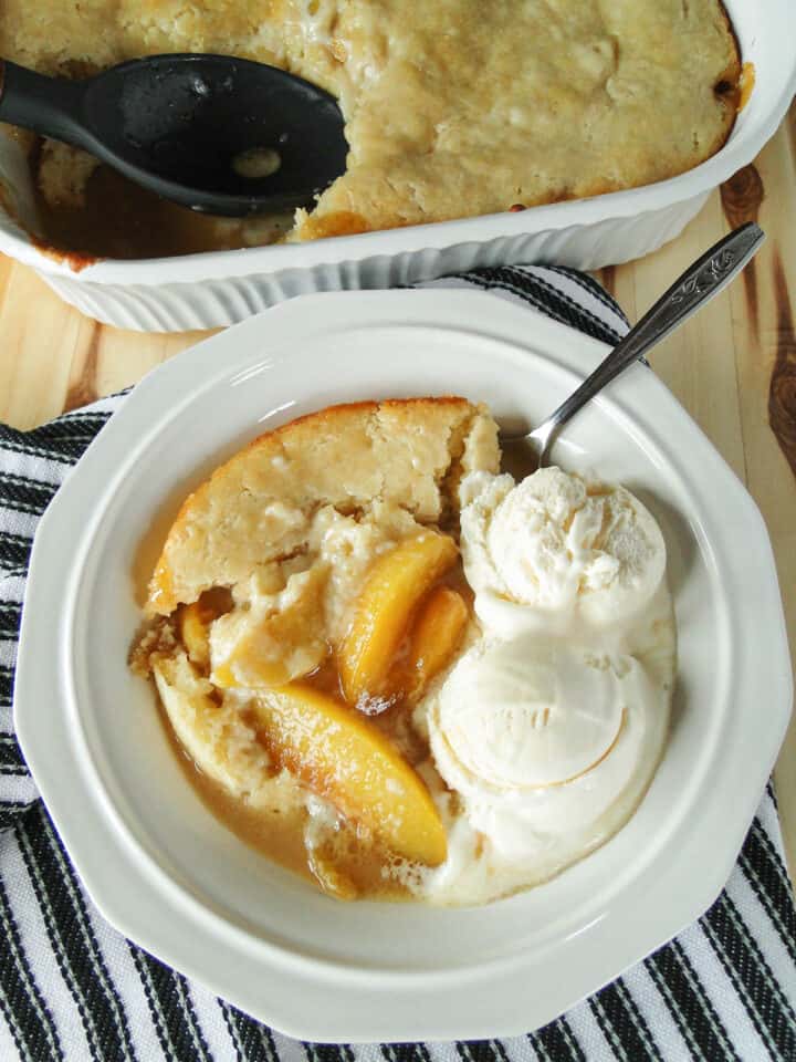 Peach cobber in white bowl with ice cream and spoon in front of baking dish of more cobbler.