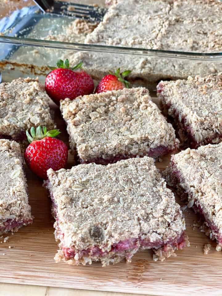Strawberry rhubarb oat bars sliced into squares and on a board in front of the dish of bars. 