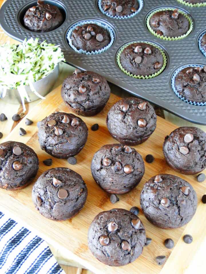 Healthy double chocolate zucchini muffins on a board in front of pan of muffins.