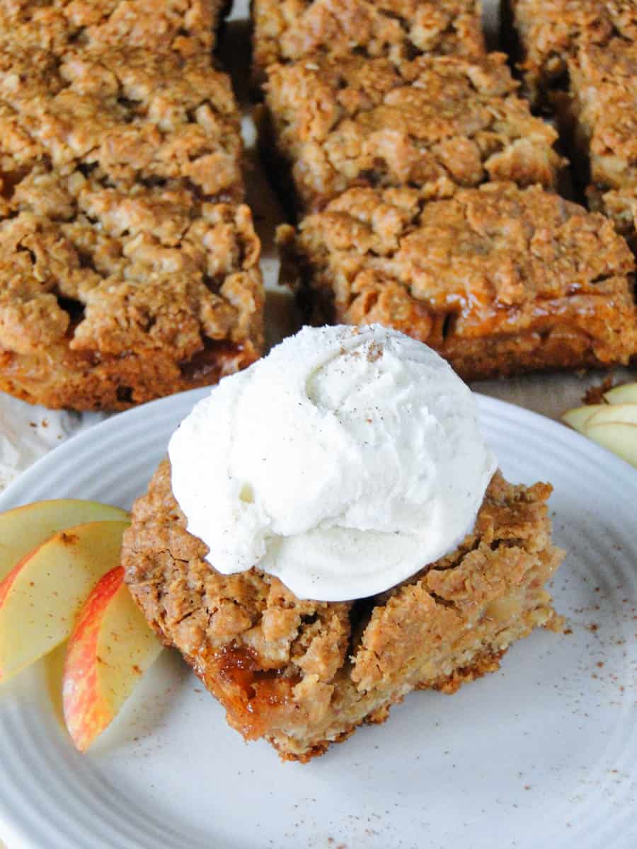 Apple pie bar with scoop of vanilla ice cream on white round plate in front of sliced bars.