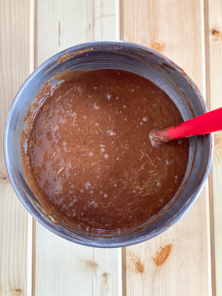 Zucchini folded into cake batter with red spatula in a mixing bowl.