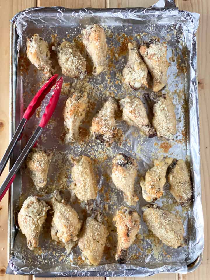 Baked chicken wings tossed with parmesan on sheet pan with tongs.