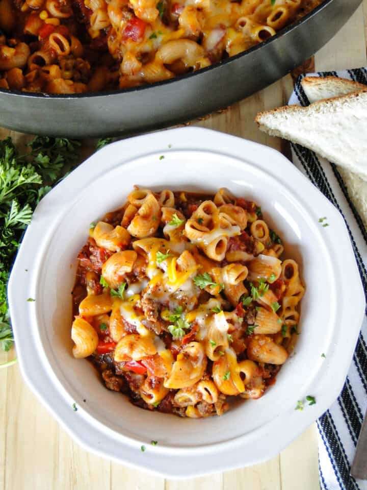 Serving of easy skillet goulash in white round bowl in front of skillet of goulash.