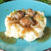 Side view of heaping mashed potatoes topped with Salisbury Steak Meatballs on blue round plate.