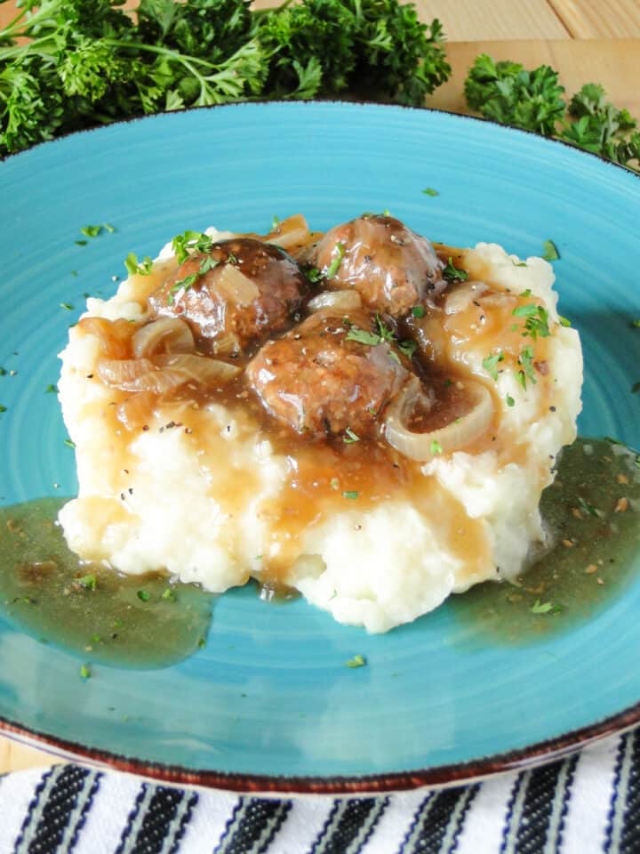 Side view of heaping mashed potatoes topped with Salisbury Steak Meatballs on blue round plate.