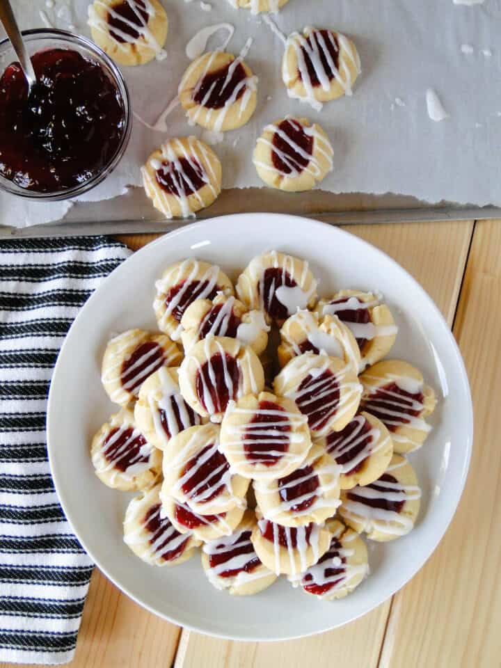 Top view of raspberry almond thumbprint cookies piled on white round plate in front of cookie sheet of cookies.
