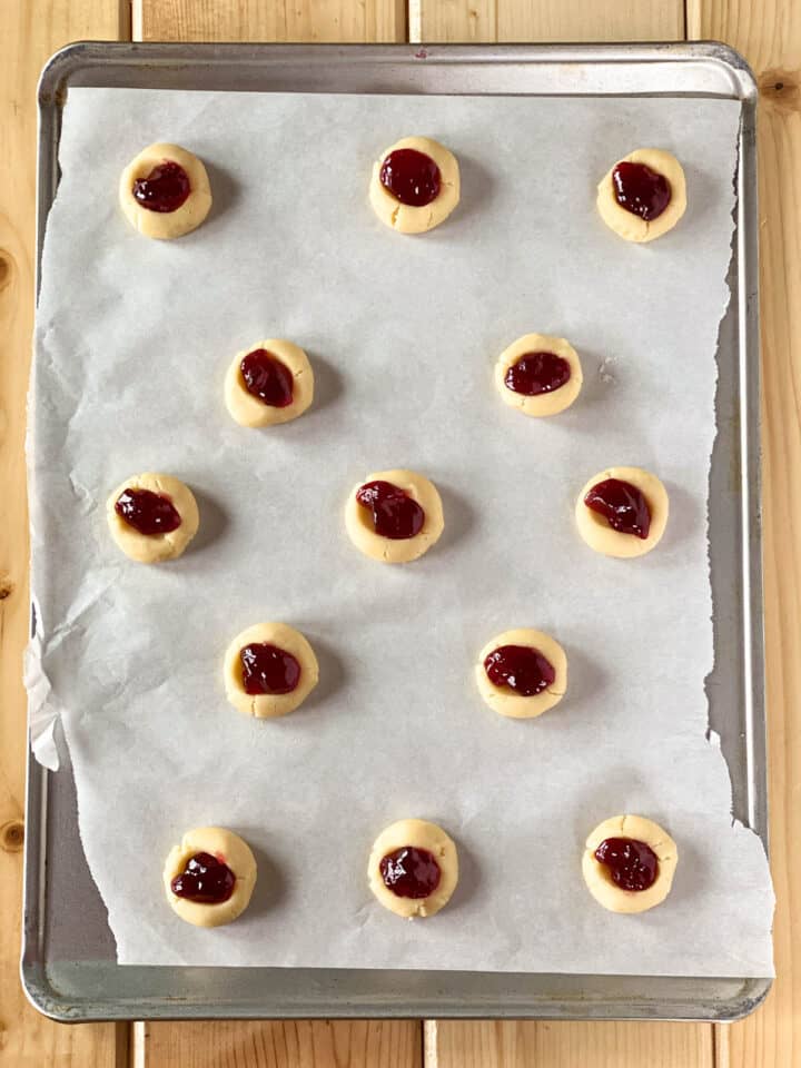 Raspberry jam added to thumbprints in cookies on cookie sheet.