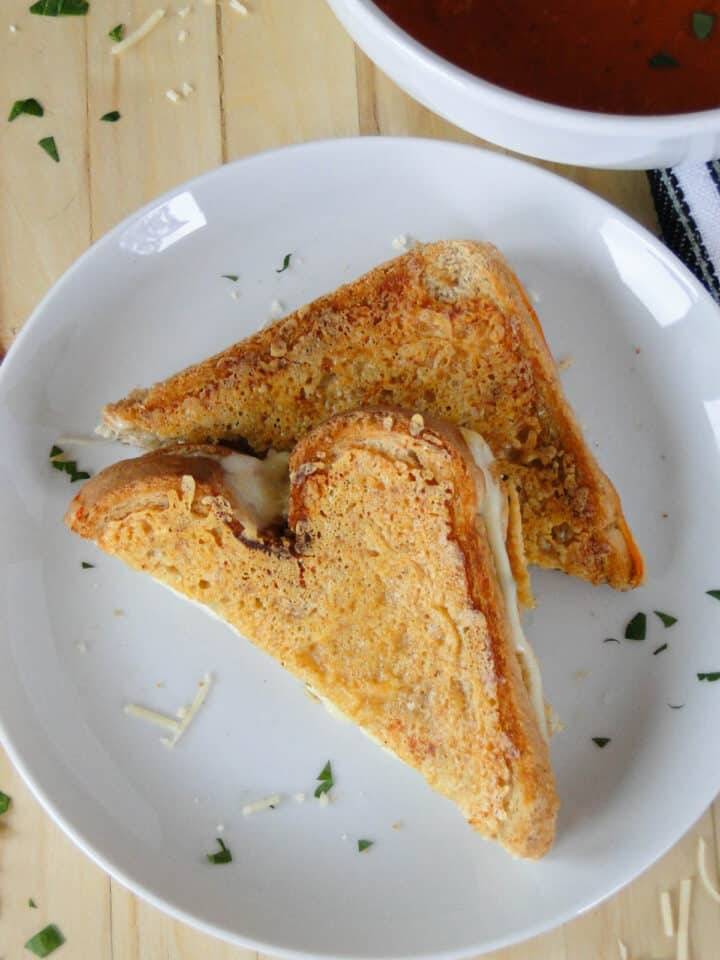 Parmesan crusted grilled cheese cut in half with one half on top of other on plate in front of bowl of tomato soup.