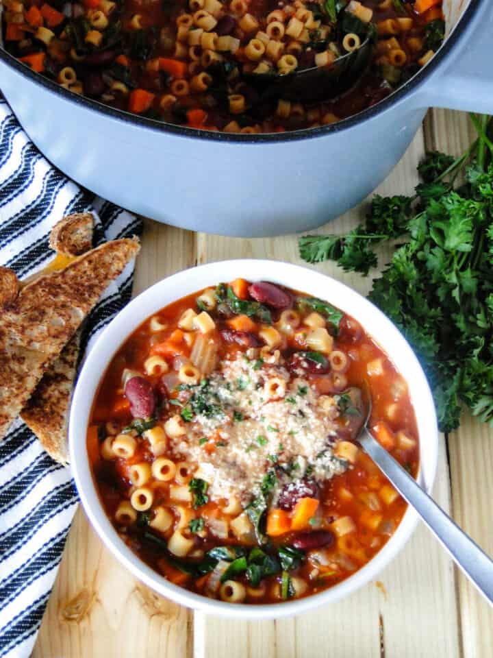 Italian minestrone soup in white round bowl with spoon in front of pot of soup.