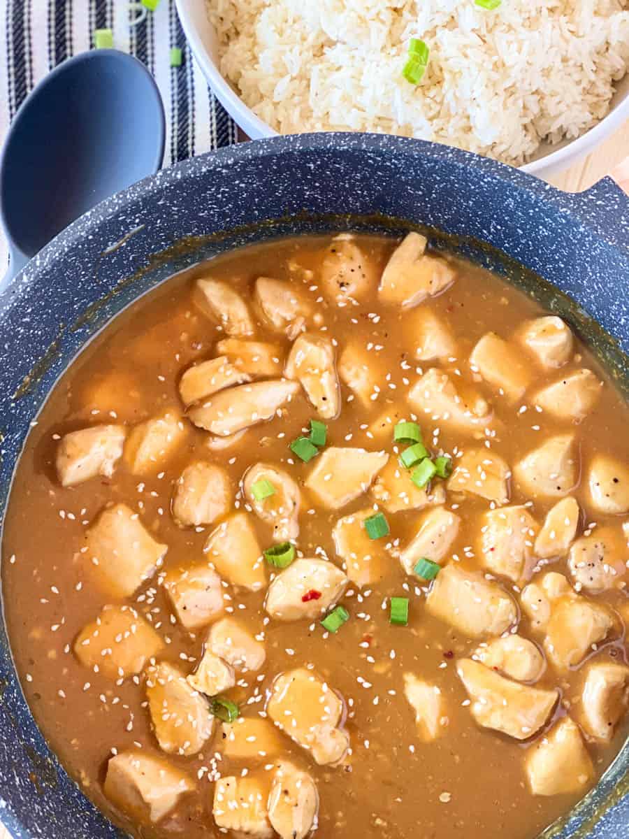 Easy orange chicken in skillet garnished with green onion and sesame seeds with bowl of rice and serving spoon on the side.