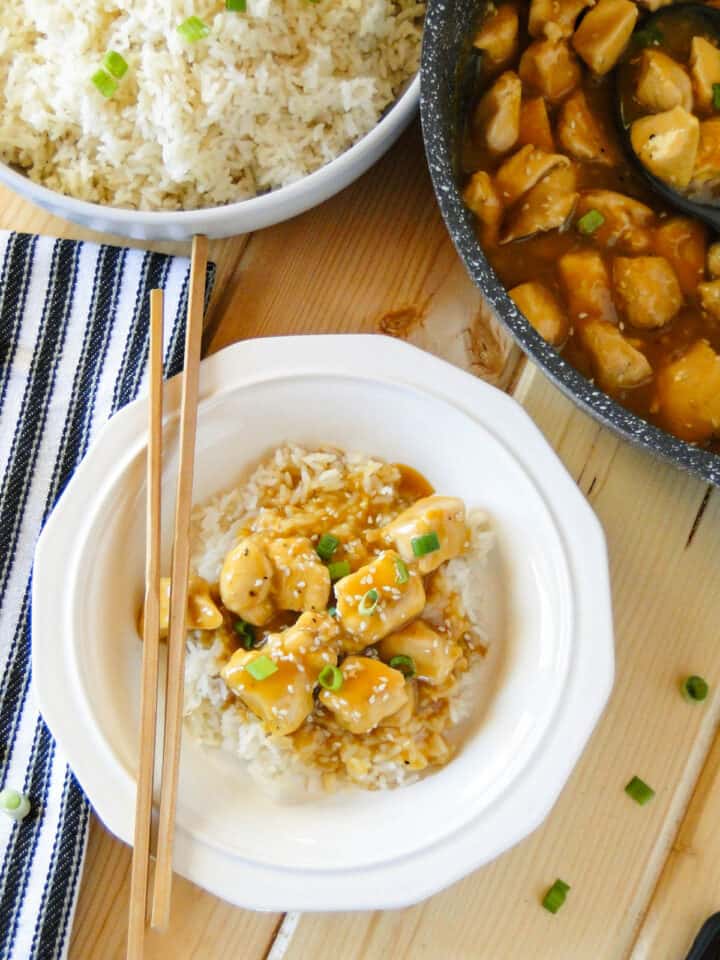 Top view of easy orange chicken over rice in white round bowl with chop sticks in front of skillet of orange chicken and bowl of white rice.